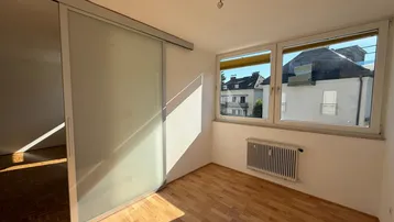 Helles Zimmer mit Parkettboden und großen Fenstern, die viel Tageslicht hereinlassen.