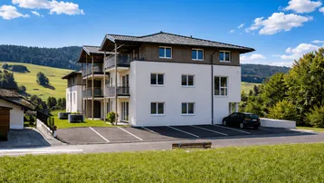 Modernes Mehrfamilienhaus mit Balkonen und Parkplätzen, eingebettet in eine grüne Hügellandschaft.