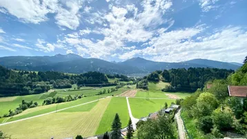 Weitläufiger Panoramablick auf die umliegende Berglandschaft mit grünen Wiesen und Wäldern unter einem blauen Himmel.