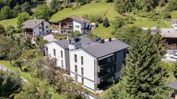 Luftaufnahme eines modernen Mehrfamilienhauses in einer grünen Berglandschaft mit Brücke im Hintergrund.