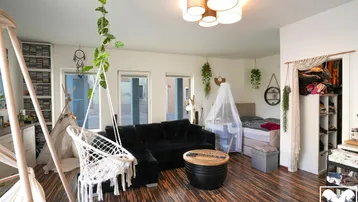 Geräumiger Wohn- und Schlafbereich mit dunklem Holzboden, gemütlicher Sitzecke, Babybett und offener Garderobe.