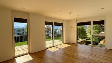 Heller, leerer Wohnbereich mit Echtholzparkett und großen Fenstern mit Zugang zur Terrasse.