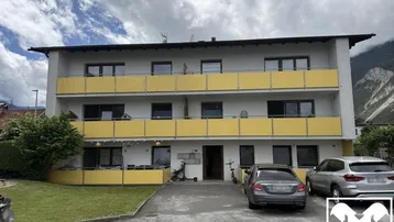 Mehrfamilienhaus mit gelben Balkonen und Parkplätzen vor alpiner Bergkulisse unter bewölktem Himmel.