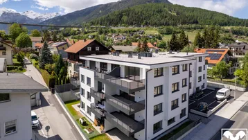 Moderne Wohnanlage mit Balkonen in alpiner Umgebung, umgeben von Grünflächen und Bergen im Hintergrund.