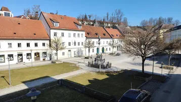 Blick auf einen historischen Stadtplatz mit traditionellen Gebäuden, Grünflächen und einem Brunnen unter blauem Himmel.
