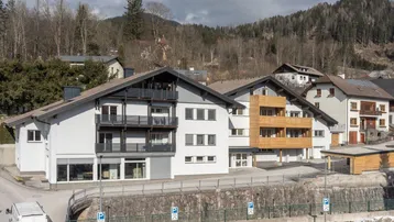 Modernes Mehrfamilienhaus mit mehreren Balkonen und Blick auf bewaldete Hügel in alpiner Umgebung.
