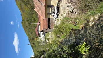 Ländliche Ansicht eines Hauses am Bachlauf mit bewaldetem Hügel im Hintergrund unter blauem Himmel.