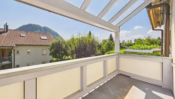 Überdachter Balkon mit Glasdach und weitem Blick auf grüne Bäume und Berge.