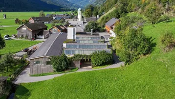 Luftaufnahme eines Gebäudekomplexes in einer ländlichen Umgebung mit grünen Hügeln und einem Dorf im Hintergrund.