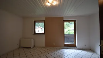 Helles Zimmer mit Fliesenboden, Balkonzugang und Blick ins Grüne durch die Fenster.