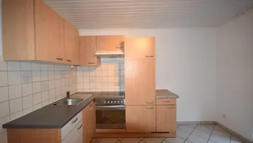 Einfache Küche mit hellen Holzschränken, dunkler Arbeitsplatte und weißen Wandfliesen.