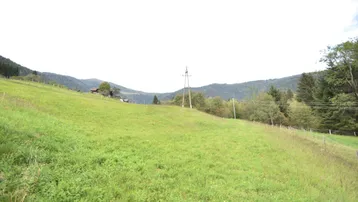Weitläufige grüne Wiesenlandschaft mit sanften Hügeln und bewaldeten Bergen im Hintergrund unter bewölktem Himmel.