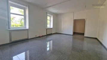 Geräumiges, leeres Wohnzimmer mit glänzendem Fliesenboden und großen Fenstern, die viel Tageslicht hereinlassen.