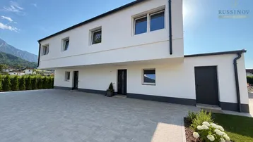 Modernes Einfamilienhaus mit weißer Fassade, Flachdach und gepflasterter Einfahrt vor Bergkulisse.