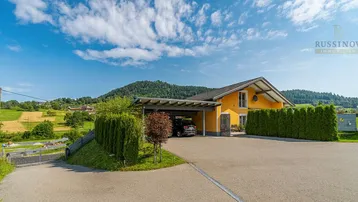 Gelbes Einfamilienhaus mit Carport und gepflegter Hecke vor einer malerischen Hügellandschaft.