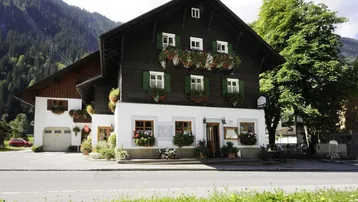 Traditionelles österreichisches Gebäude mit dunkler Holzfassade, weißen Wänden und Blumenkästen vor einer Bergkulisse.