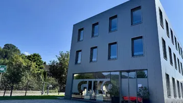 Modernes Bürogebäude mit grauer Fassade und großen Glasfenstern im Eingangsbereich unter blauem Himmel.