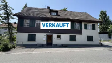 Zweistöckiges Wohnhaus mit Satteldach, grünen Fensterläden und weißer Fassade an einer Straße.