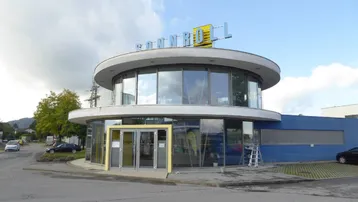 Modernes, rundes Gebäude mit großer Glasfassade und umliegenden Parkplätzen unter bewölktem Himmel.