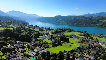 Panoramablick auf eine Siedlung am Ufer eines Sees, umgeben von grünen Hügeln und Bergen unter blauem Himmel.
