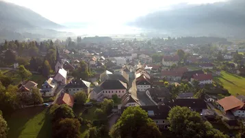 Luftaufnahme eines malerischen Dorfes in einem Tal, umgeben von grünen Hügeln und leichtem Morgennebel.