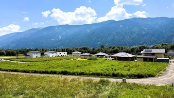 Weitläufige Landschaft mit grünen Wiesen, modernen Häusern und majestätischen Bergen im Hintergrund unter blauem Himmel.