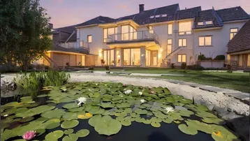Großzügiges Anwesen mit Teich und beleuchteter Fassade bei Sonnenuntergang, umgeben von Grünflächen.