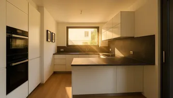 Moderne Küche mit weißen Schränken, dunklen Arbeitsplatten und eingebauten Geräten, hell beleuchtet durch ein Fenster.
