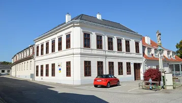 Weißes historisches Gebäude mit braunen Fenstern und einem roten Auto davor auf einem gepflasterten Platz.