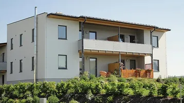 Modernes Mehrfamilienhaus mit weißer Fassade, Balkonen und einer möblierten Terrasse im Grünen.