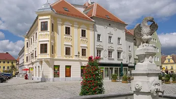 Historisches Gebäude an einem belebten Stadtplatz mit einem dekorativen Brunnen und Blumenarrangement.