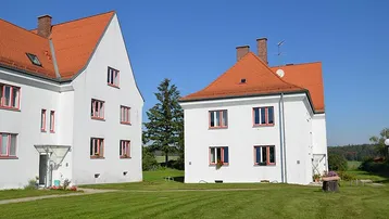 Zwei traditionelle weiße Gebäude mit roten Dächern, umgeben von einer weitläufigen grünen Wiese.