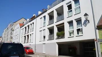 Moderne Wohngebäude mit Balkonen und Tiefgaragenzufahrt an einer sonnigen Straße.