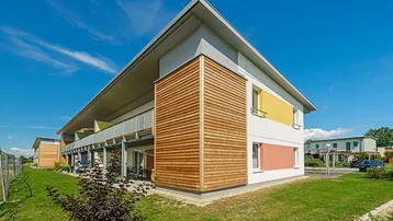 Modernes Mehrfamilienhaus mit farbigen Akzenten, Holzverkleidung und gepflegter Grünfläche unter blauem Himmel.