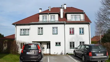 Weißes Mehrfamilienhaus mit rotem Ziegeldach und Parkplätzen vor dem Gebäude.