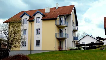 Mehrfamilienhaus mit gelber Fassade, roten Ziegeldächern und kleinen Balkonen in ländlicher Umgebung.