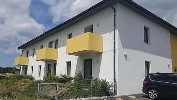 Modernes Mehrfamilienhaus mit weißer Fassade, gelben Balkonen und Satteldach unter blauem Himmel.