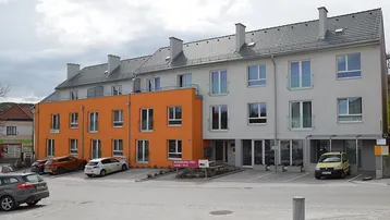 Ansicht des modernen Mehrfamilienhauses mit orangefarbenem Akzent und Parkplätzen im Freien vor einer grünen Hügellandschaft.