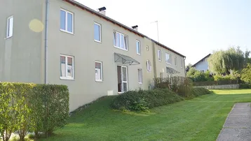 Grünes Mehrfamilienhaus mit gepflegtem Rasen und Hecke in ländlicher Umgebung.