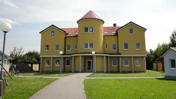 Gelbes Mehrfamilienhaus mit markantem Rundturm und rotem Ziegeldach in grüner Umgebung.
