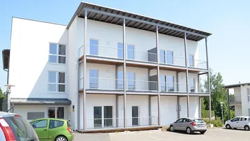 Modernes Mehrfamilienhaus mit mehreren Balkonen und Außenparkplätzen unter blauem Himmel.