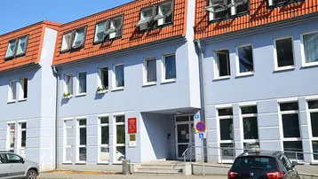 Ansicht eines Bürogebäudes mit hellblauer Fassade, roten Dachziegeln und einer Rollstuhlrampe am Eingang.