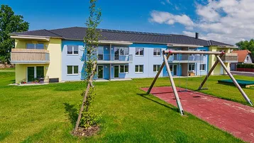 Mehrfamilienhaus mit farbenfroher Fassade, Balkonen und einem Spielplatz im Grünen.