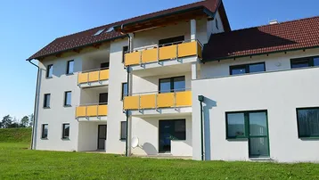 Modernes Mehrfamilienhaus mit weißer Fassade, gelben Balkonen und rotem Ziegeldach, umgeben von Grünflächen.