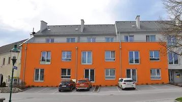Modernes Mehrfamilienhaus mit leuchtend oranger Fassade und Parkplätzen vor dem Gebäude.