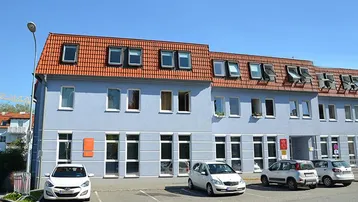 Ansicht der modernen Gebäudefassade mit blauer Farbe, roten Ziegeldächern und zahlreichen Dachfenstern sowie Parkplätzen davor.