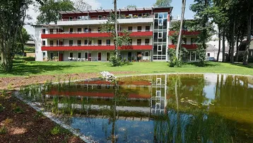 Modernes Mehrfamilienhaus mit roten Akzenten, großzügigen Balkonen und einem Teich in einer grünen Umgebung.