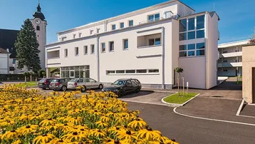 Modernes weißes Gebäude mit gelben Blumen im Vordergrund und einem Kirchturm im Hintergrund.