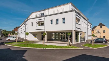 Modernes weißes Gebäude mit großen Fenstern und Balkonen unter blauem Himmel an einem sonnigen Tag.