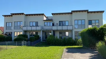 Modernes Mehrfamilienhaus mit heller Fassade, Balkonen und gepflegtem Vorgarten unter blauem Himmel.